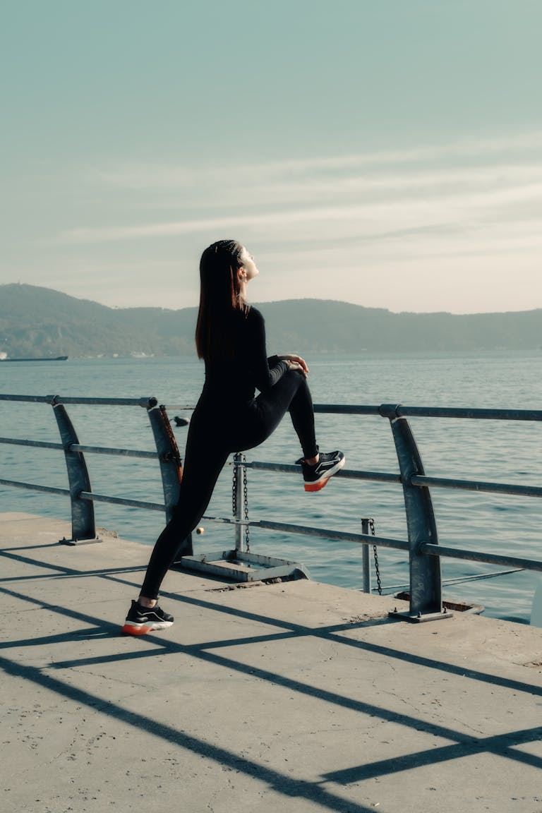 Woman in Sportswear Stretching Her Legs against the Railing along the Shore